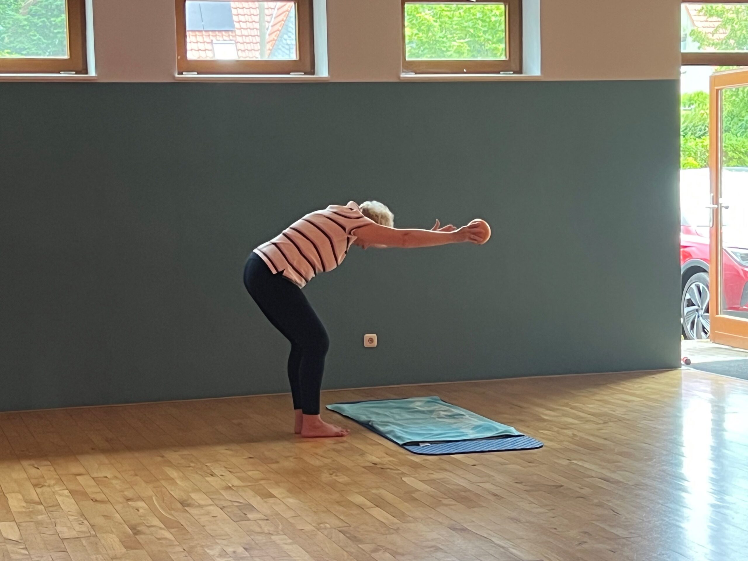 Teilnehmerin steht gebeugt und lehnt sich mit dem Rücken nach vorne, ihre Arme streckt sie nach vorne in der rechen Hand hält sie einen kleinen Ball.